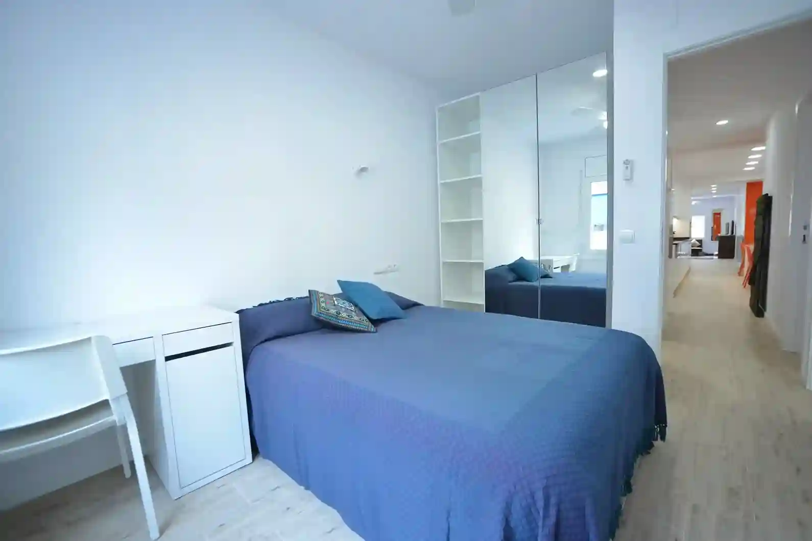 Vista lateral de la habitación con cama doble, escritorio, armario con puertas de espejo y estantería